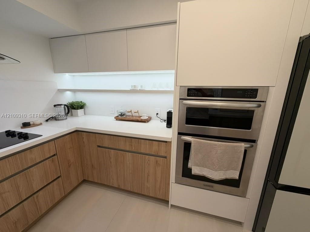 Kitchen featuring two-tone cabinetry with wood-finish lower units, white upper cabinets, white solid-surface countertops, integrated under-cabinet lighting, and a stainless steel wall oven