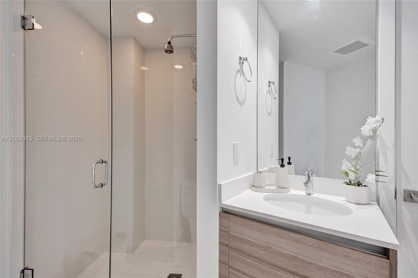 Bathroom featuring a wood-finish vanity with a white countertop and oval undermount sink, large wall-mounted mirror, and glass-enclosed shower with tiled walls