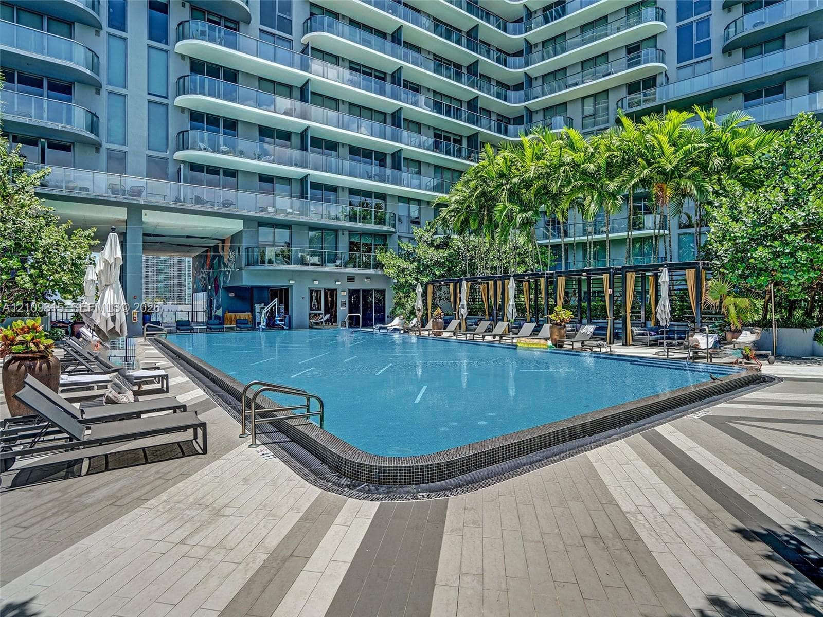 Swimming pool with tile surround, multi-level building exterior with glass-paneled balconies, cabana structure, and paved deck