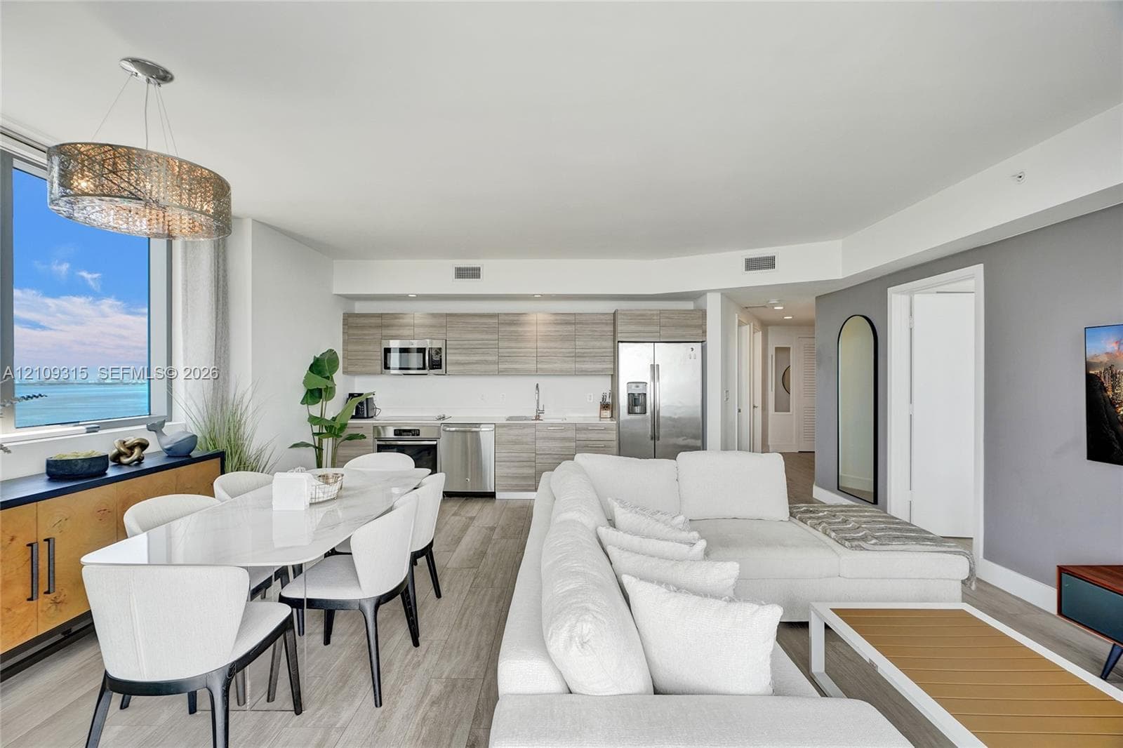 Open-concept living space featuring wood-finish flooring, floor-to-ceiling windows, modern kitchen cabinetry, stainless steel appliances, and a contemporary drum pendant light fixture