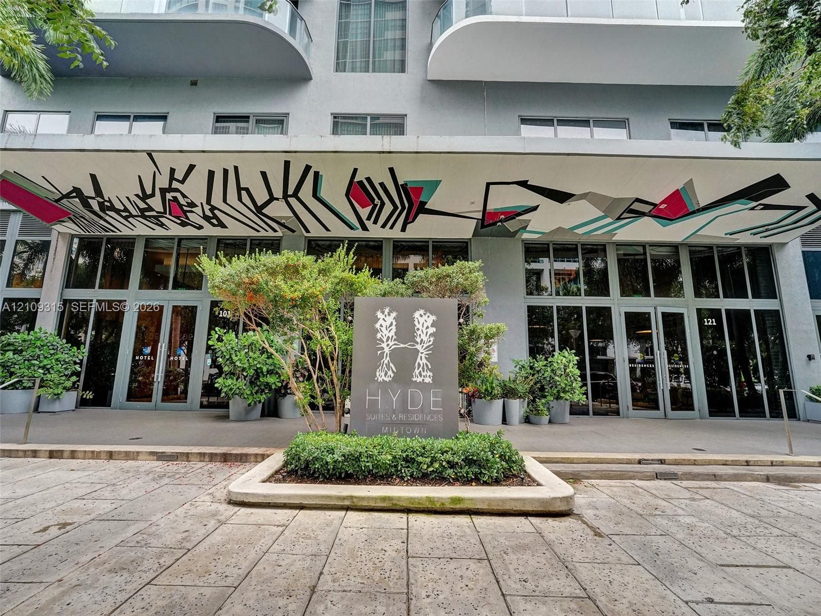 Exterior entrance featuring floor-to-ceiling glass paneling, a concrete canopy with artistic mural, paved stone walkway, and structured landscaping