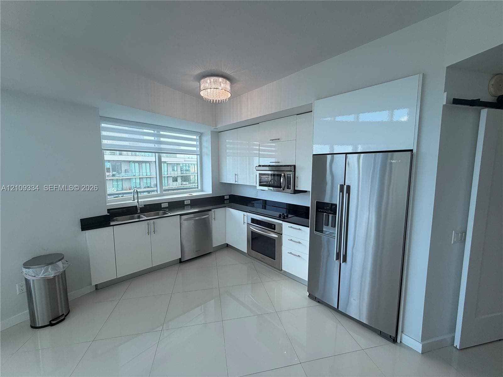 Kitchen featuring high-gloss white cabinetry, stainless steel appliances, dark stone countertops, polished tile flooring, and a crystal flush-mount light fixture