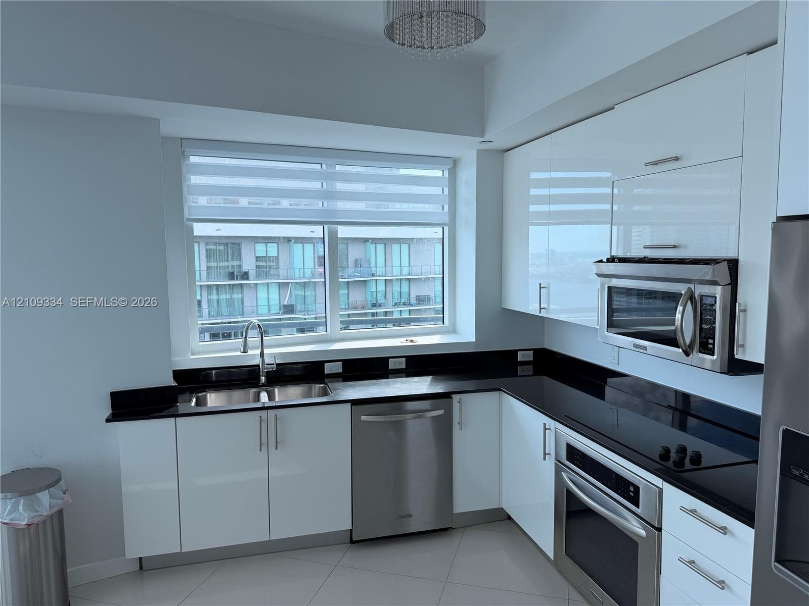 Kitchen featuring high-gloss white cabinetry, dark stone-finish countertops, stainless steel appliances, large double-basin under-mount sink, and a modern drum-style ceiling fixture