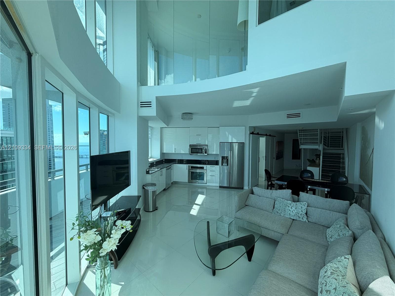 Double-height living area featuring floor-to-ceiling glass windows, high-gloss kitchen cabinetry, stainless steel appliances, and interior glass mezzanine railing