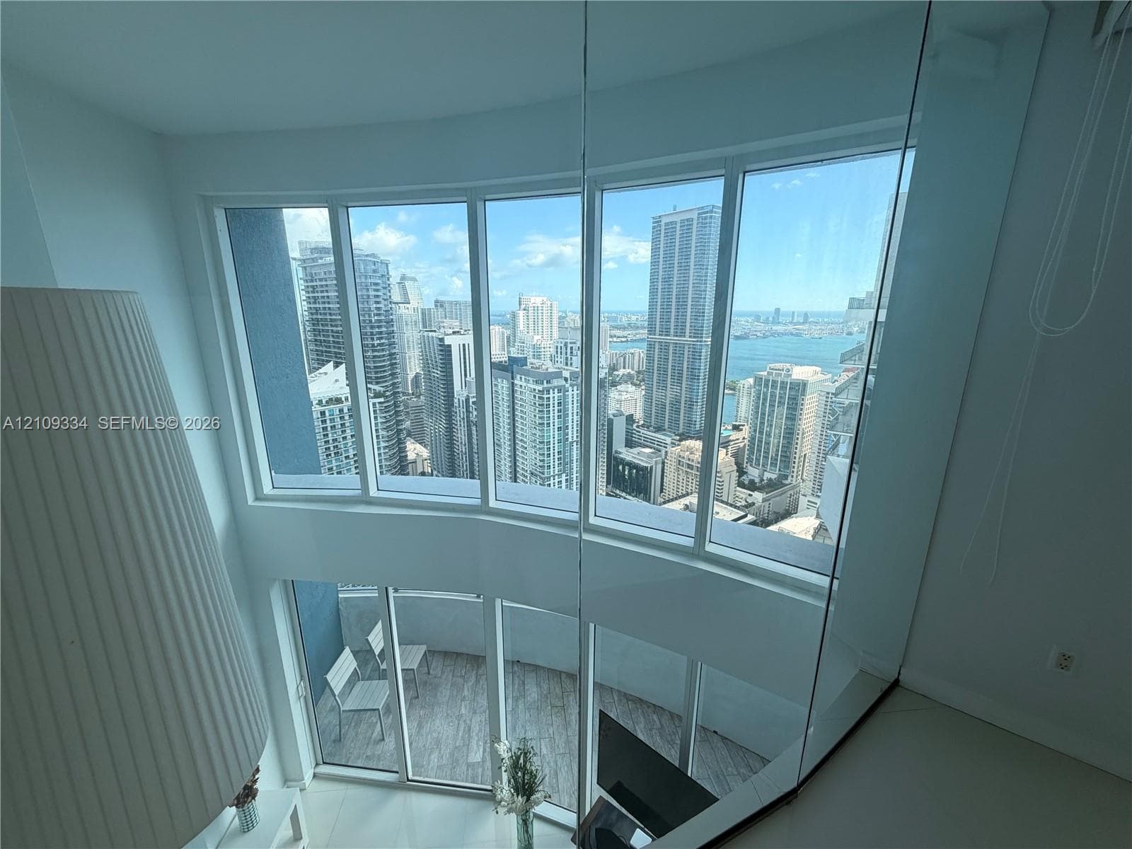 Double-height floor-to-ceiling windows featuring city and water scenery, glass partition railing, and private balcony with wood-finish flooring