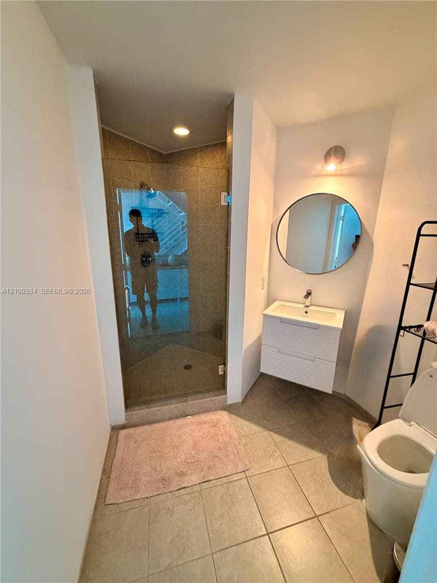 Bathroom featuring a glass-enclosed shower with tiled surround, wall-mounted white vanity with a rectangular sink, circular mirror, and large-format floor tiling