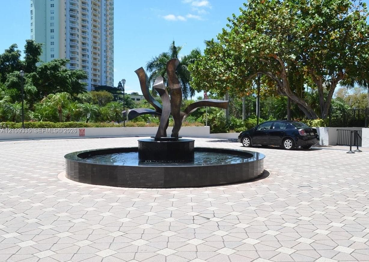 Circular stone water feature with a central metal sculptural element, set within a paved stone courtyard featuring mature landscaping