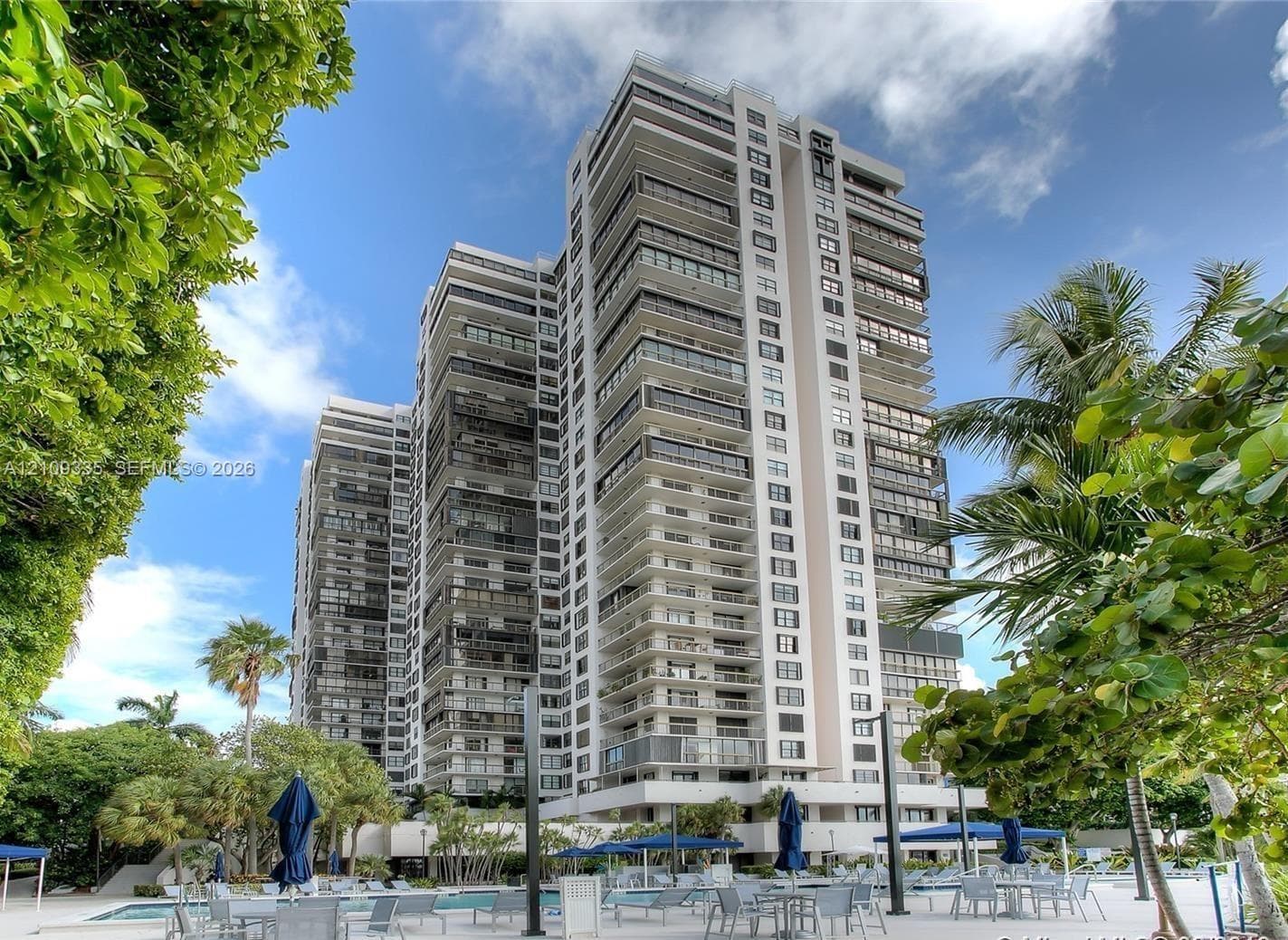 High-rise residential building featuring expansive private balconies with glass railings, concrete structural exterior, and ground-level pool deck with mature tropical landscaping
