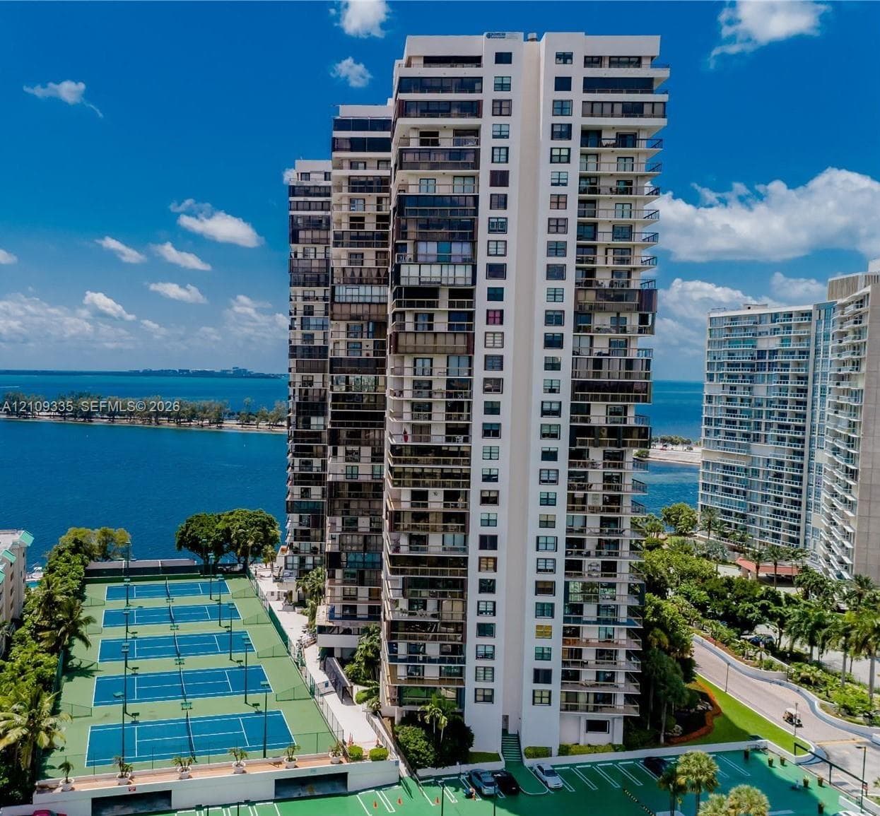 High-rise tower featuring multiple balconies, ground-level tennis courts, and immediate proximity to water