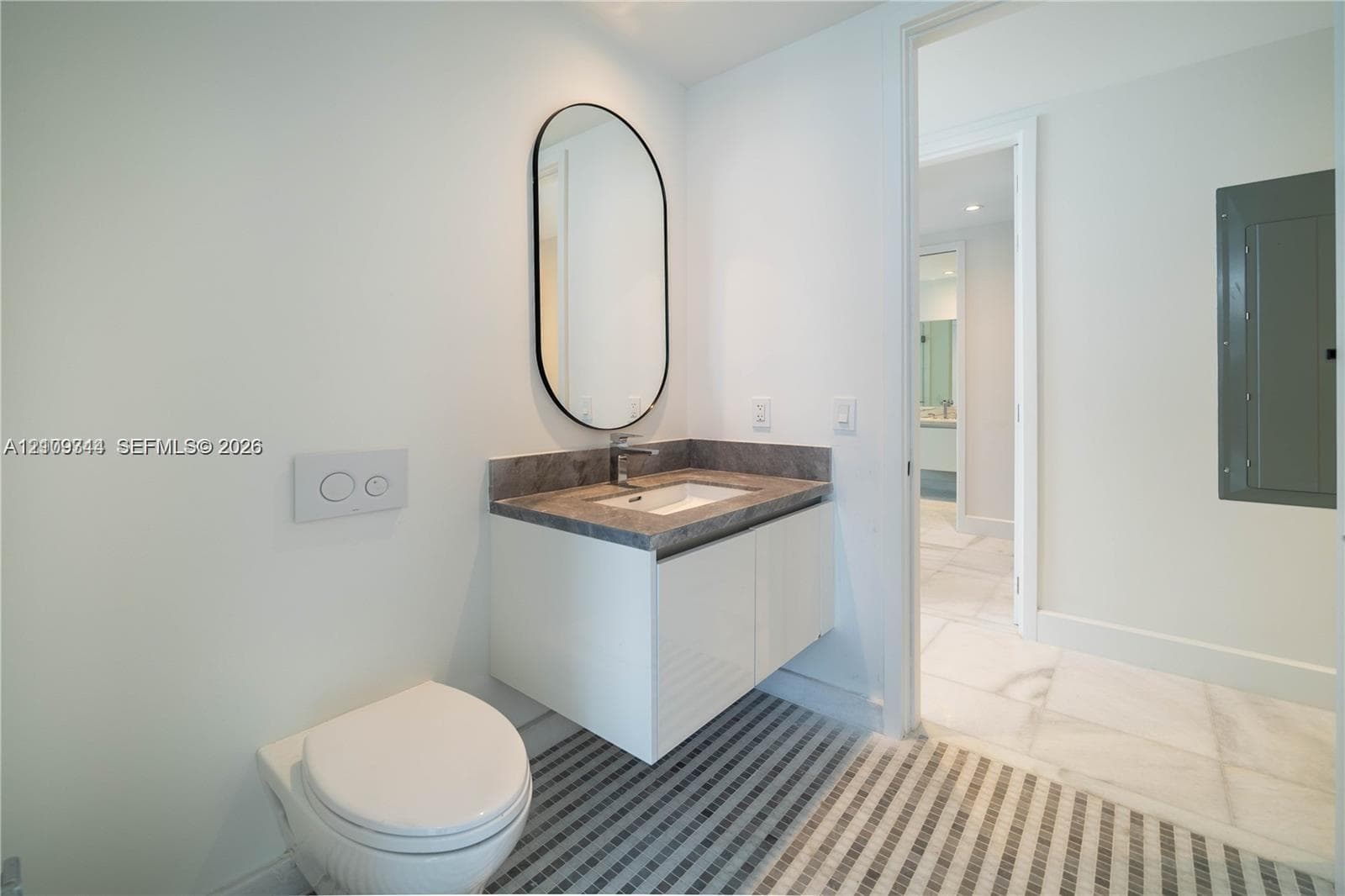 Wall-mounted white vanity with a stone countertop, integrated sink, and an arched-frame mirror, complemented by a wall-mounted toilet and multi-toned linear tiled flooring