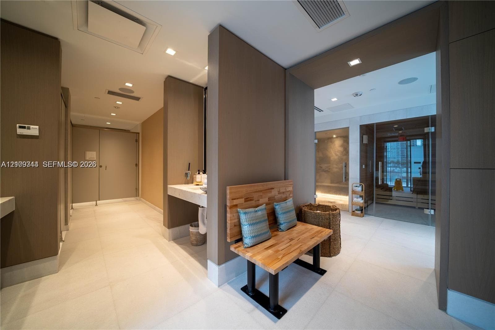 Spa-inspired facility featuring large-format porcelain tile flooring, wall-mounted stone-surface vanities, recessed ceiling lighting, and a glass-enclosed sauna room