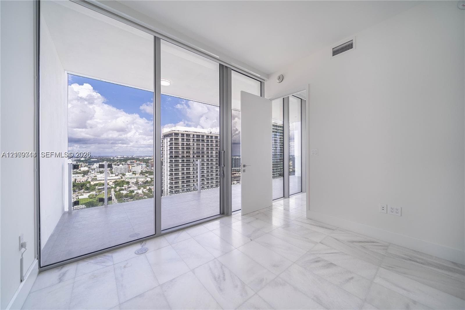 Floor-to-ceiling sliding glass doors, private balcony with metal railing, light-toned stone-finish flooring, and recessed ceiling lighting