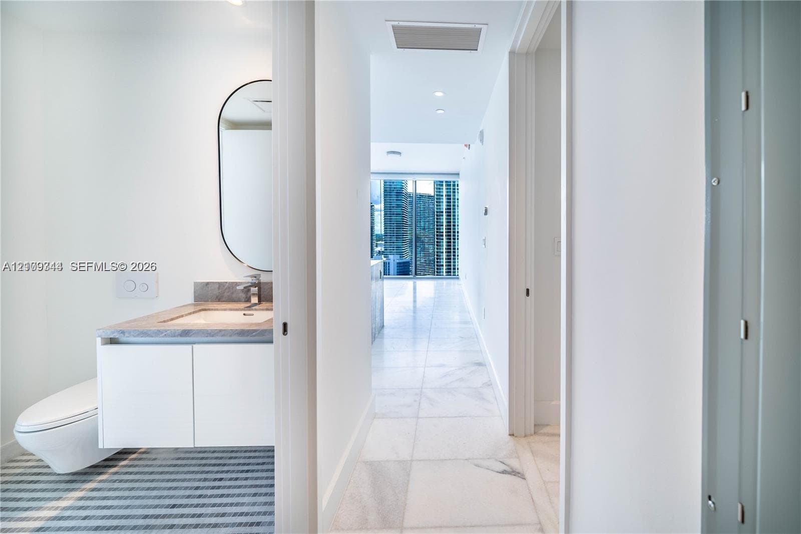 Bathroom featuring a wall-mounted toilet, vanity with a stone countertop, oval mirror, and large-format tile flooring extending into the hallway