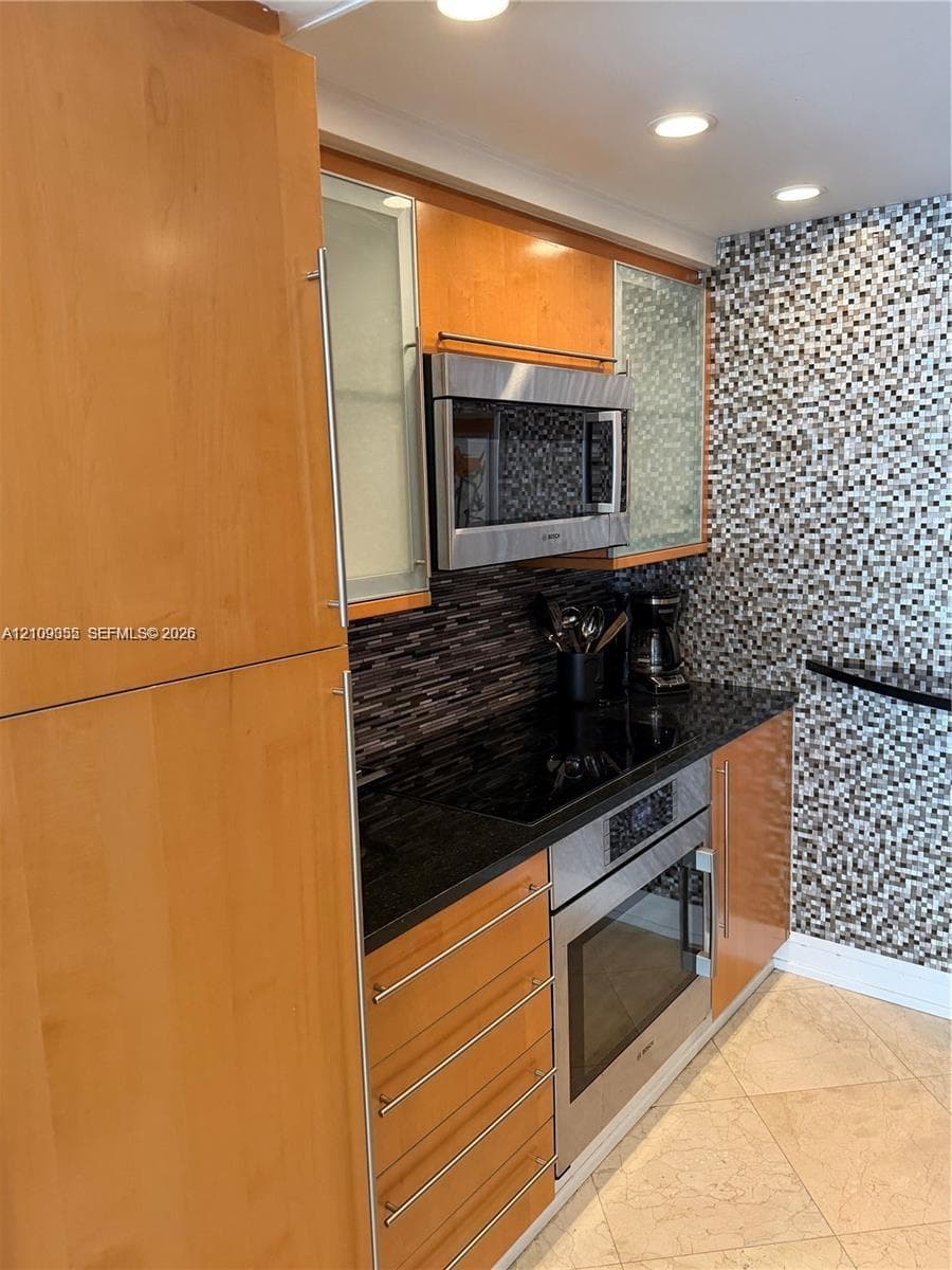 Flat-panel wood cabinetry with frosted glass inserts, stainless steel built-in microwave, integrated electric cooktop, stainless steel oven, and mosaic tile backsplash