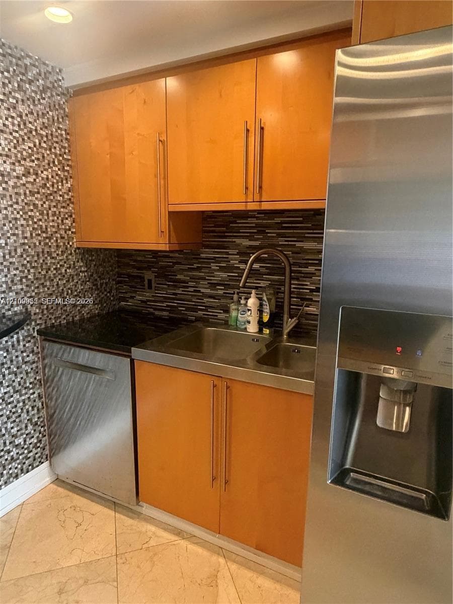 Kitchen area featuring flat-panel wood cabinetry, a stainless steel double-basin sink with a gooseneck faucet, a stainless steel dishwasher, a full-size stainless steel refrigerator, and a decorative tile backsplash