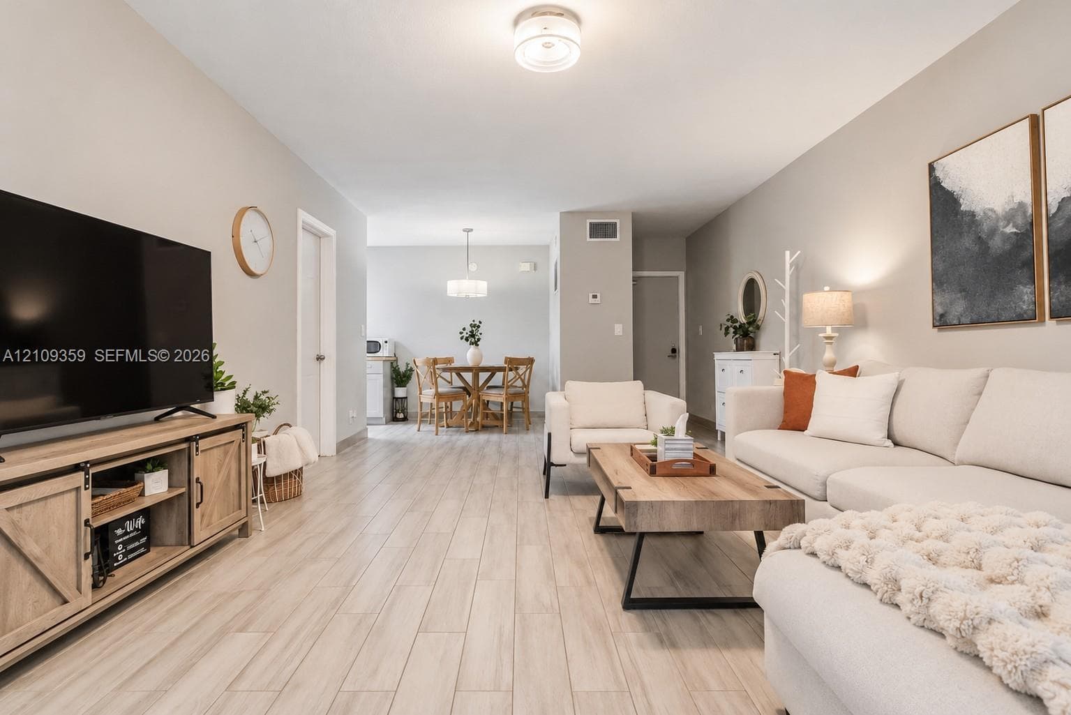 Open-concept interior featuring light wood-finish flooring, neutral wall paint, a circular brushed-nickel dining area chandelier, and a flush-mount overhead light fixture
