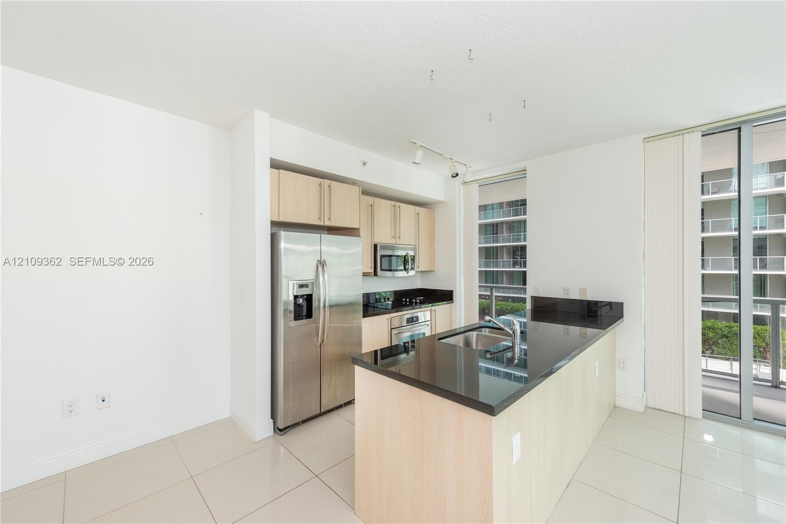 Kitchen featuring light wood-finish cabinetry, dark solid-surface countertops, stainless steel refrigerator, built-in microwave, and large-format tile flooring