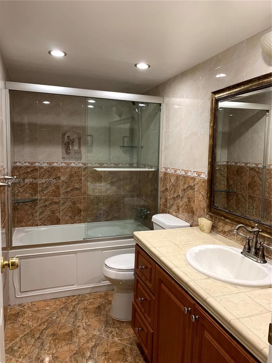 Full-bathroom featuring a tiled tub enclosure with sliding glass doors, wooden cabinetry with a tiled countertop, recessed lighting, and an integrated wall mirror