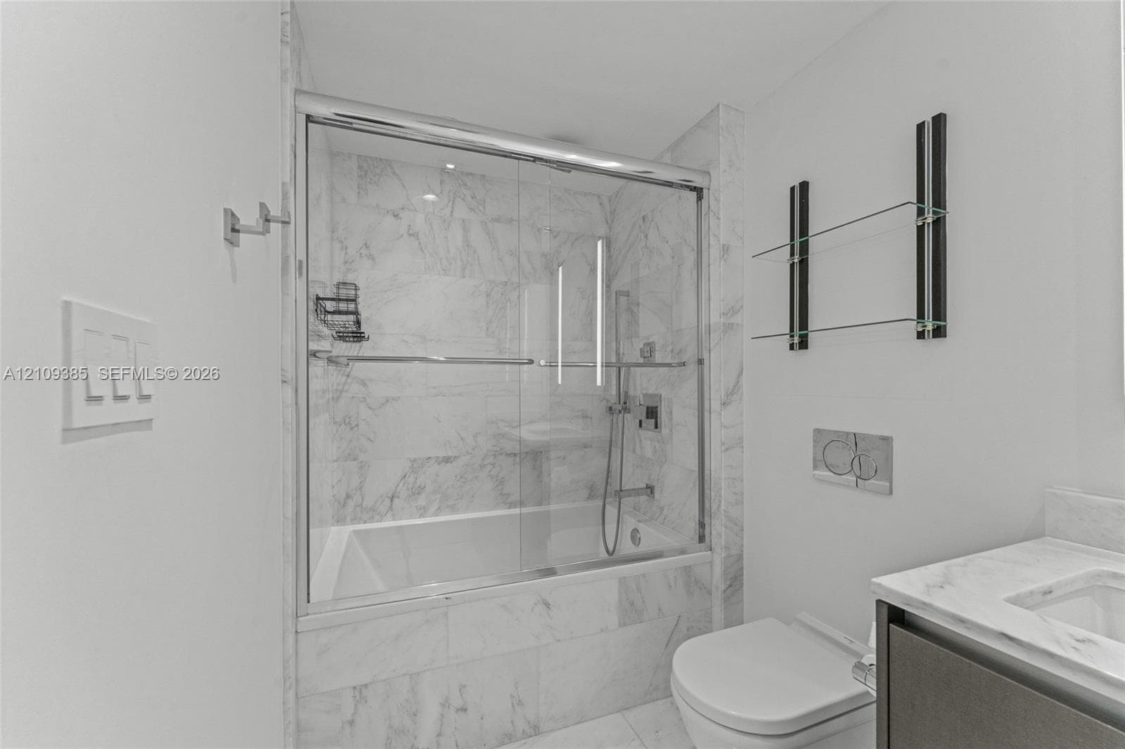 Bathroom featuring a tub-shower combination with glass sliding doors, floor-to-ceiling stone-look tiling, a wall-mounted toilet with a dual-flush actuator, built-in glass shelving, and a stone-topped vanity