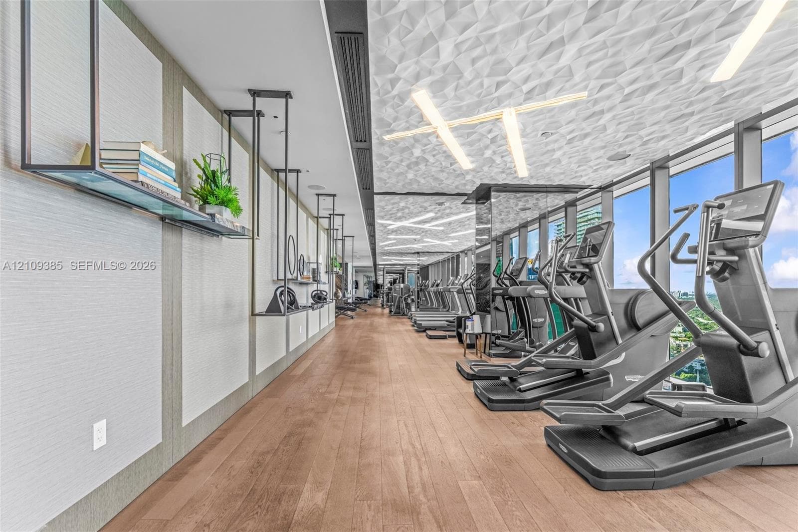 Fitness center featuring wood-finish flooring, sculptural geometric ceiling panels, linear recessed lighting, floor-to-ceiling windows, and wall-mounted metal shelving