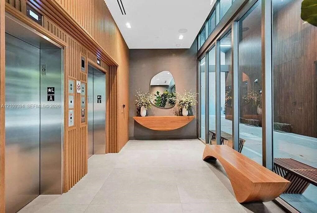 Lobby area featuring stainless steel elevator doors, floor-to-ceiling glass paneling, wood-slat wall paneling, large-format tile flooring, and a built-in wood console