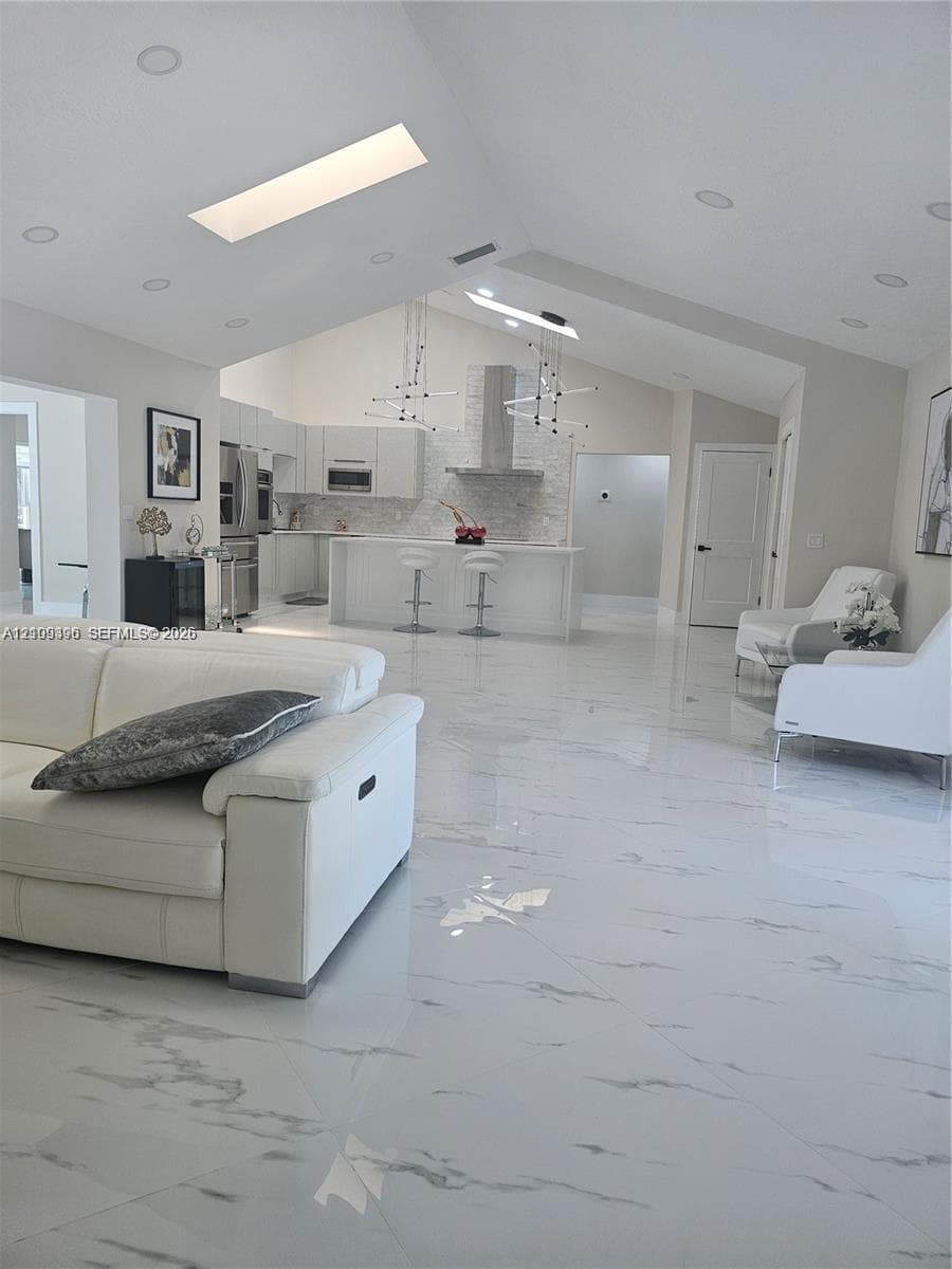 Open-concept interior featuring vaulted ceilings, skylights, large-format tile flooring, recessed lighting, and a kitchen island with a stone-finish backsplash