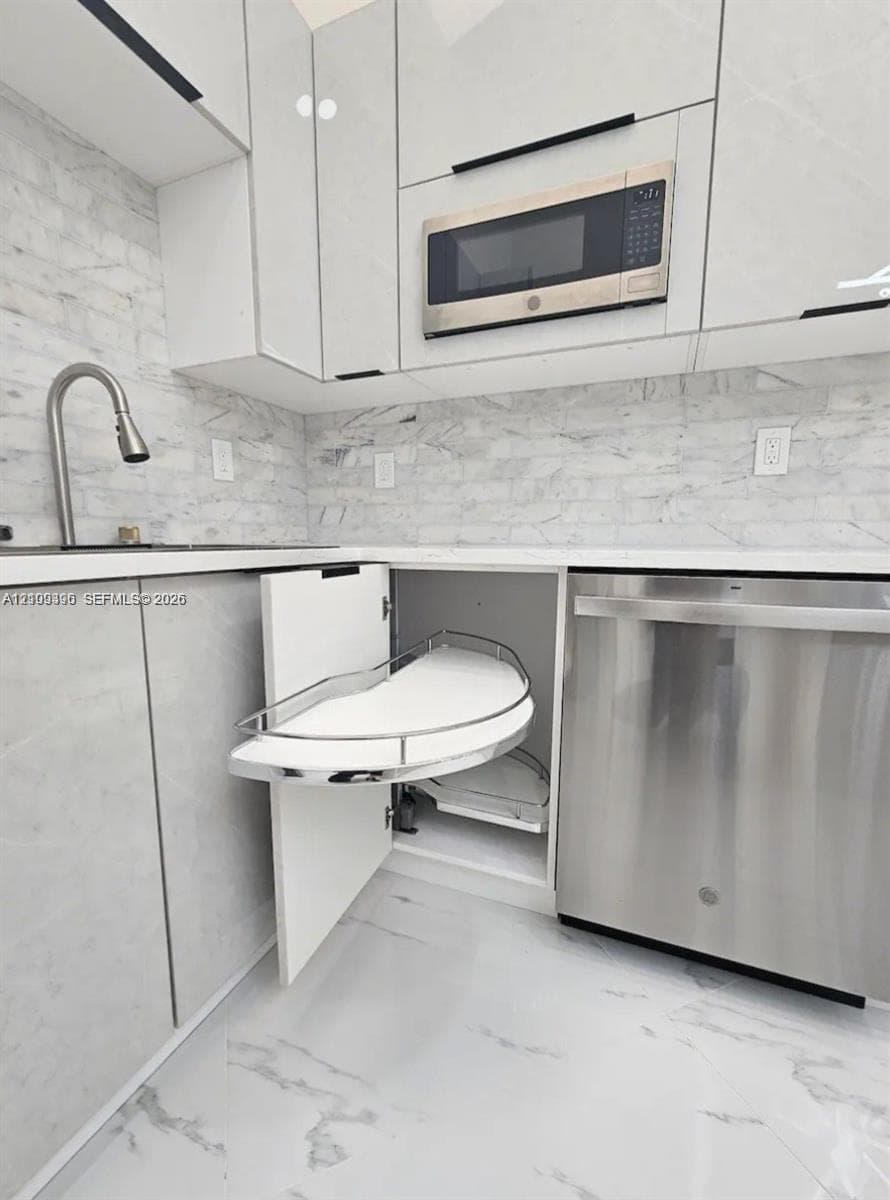 Modern kitchen featuring white flat-panel cabinetry, pull-out corner shelving, stainless steel appliances, stone-patterned backsplash, and matching stone-patterned flooring