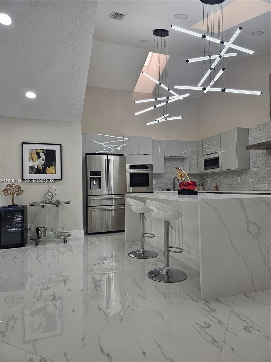 Modern kitchen featuring a waterfall island with stone-finish countertops, high-gloss cabinetry, stainless steel appliances, large-format tile flooring, and geometric multi-pendant lighting