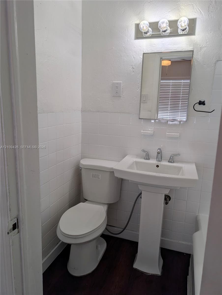 White pedestal sink featuring a chrome faucet, wall-mounted rectangular mirror, white square tile wainscoting, and wood-finish flooring