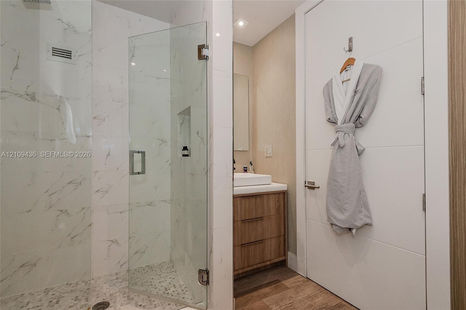 Walk-in shower featuring glass paneling, marble-style tile walls, and a pebble-stone floor, paired with a wood-finish vanity, vessel sink, and solid-core door