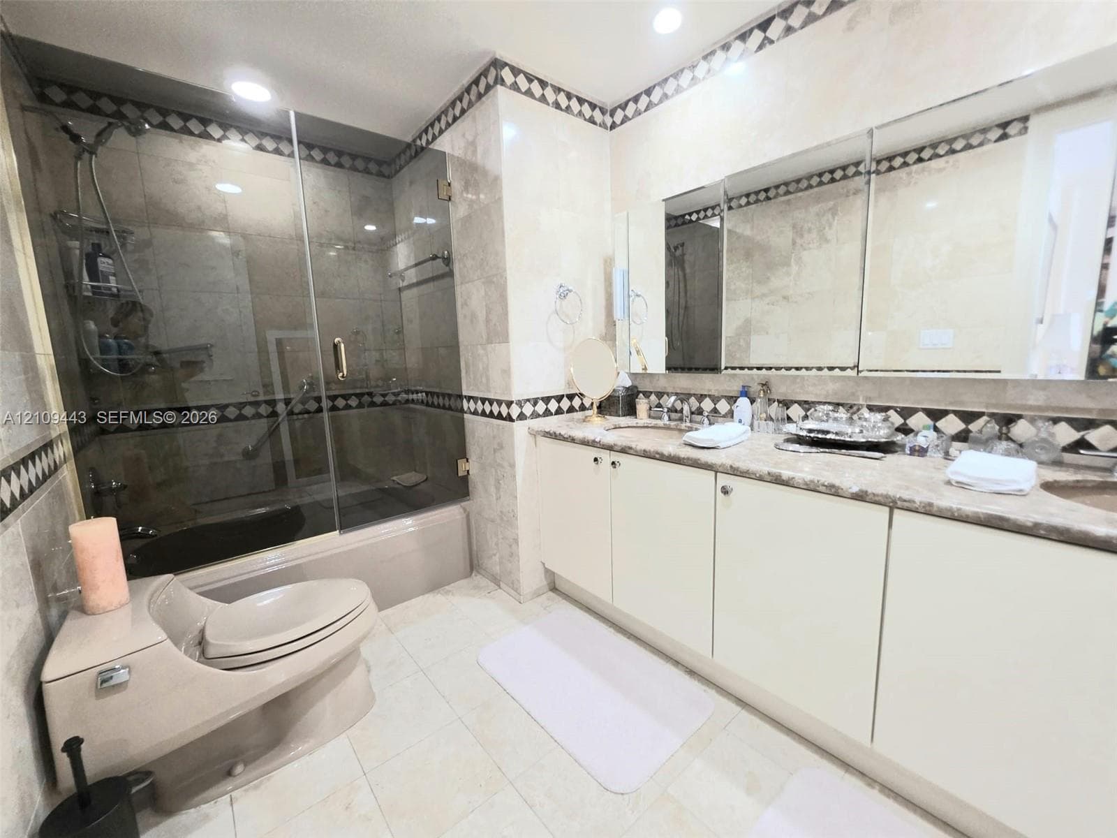 Bathroom featuring stone-tiled walls with decorative mosaic trim, dual-sink vanity with solid-surface countertop, frameless glass shower enclosure, and recessed lighting