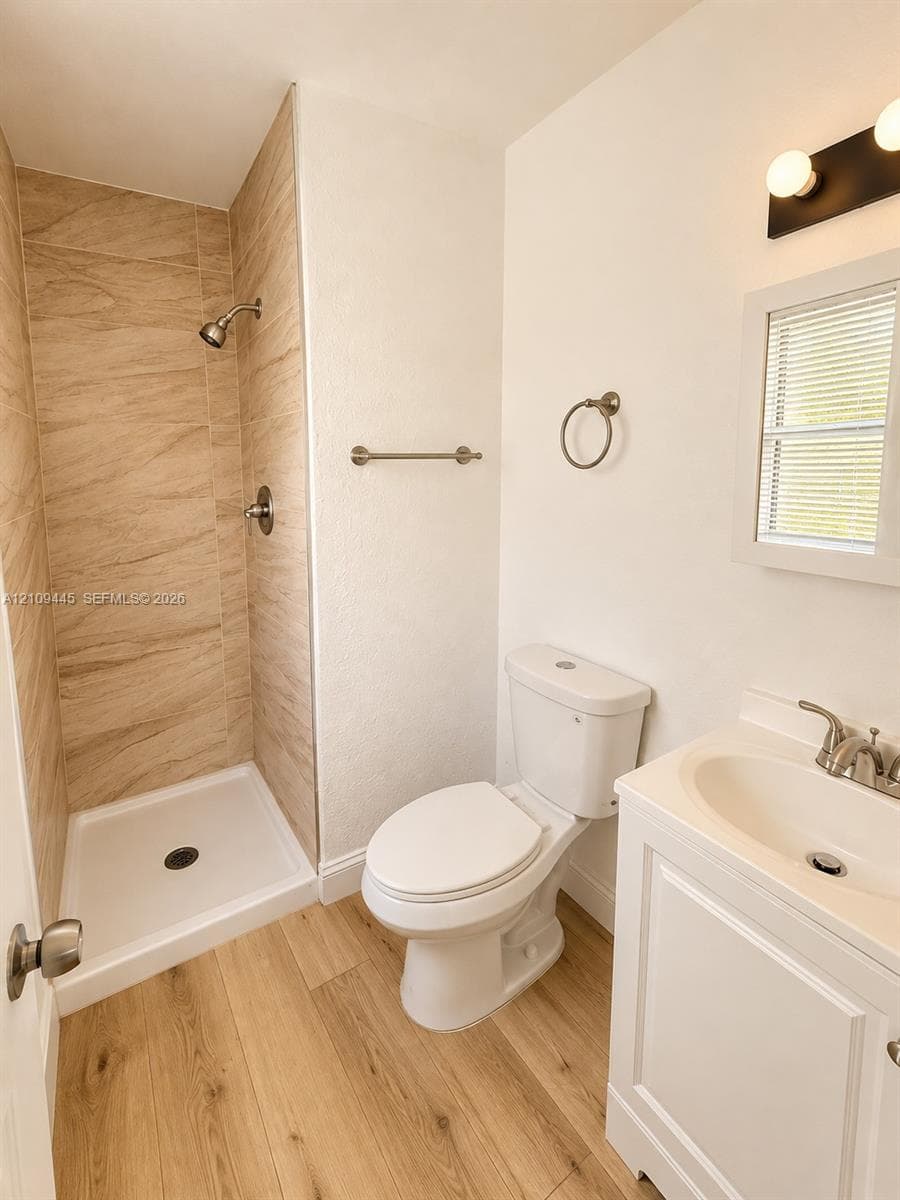 Bathroom featuring a walk-in shower with stone-finish tile surround, wood-finish flooring, white vanity cabinet with an integrated basin, and brushed nickel hardware