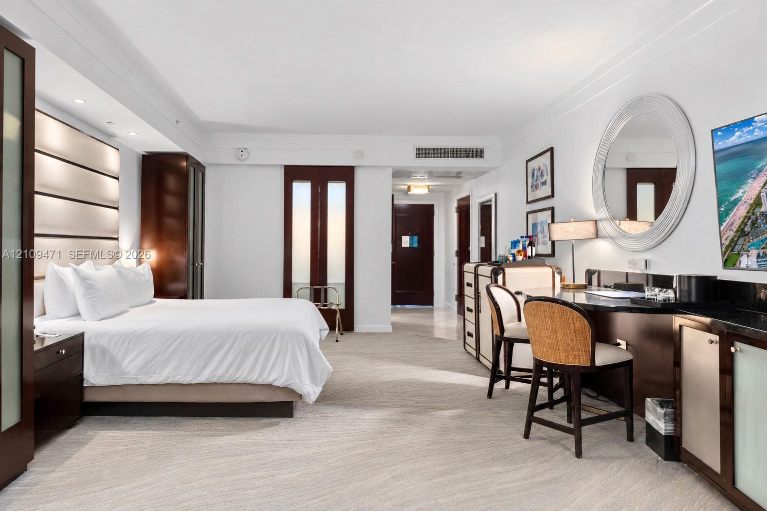 Spacious room featuring a wall-mounted panel headboard, dark wood-finish cabinetry, a built-in stone countertop desk, a circular wall mirror, and neutral-toned carpeting