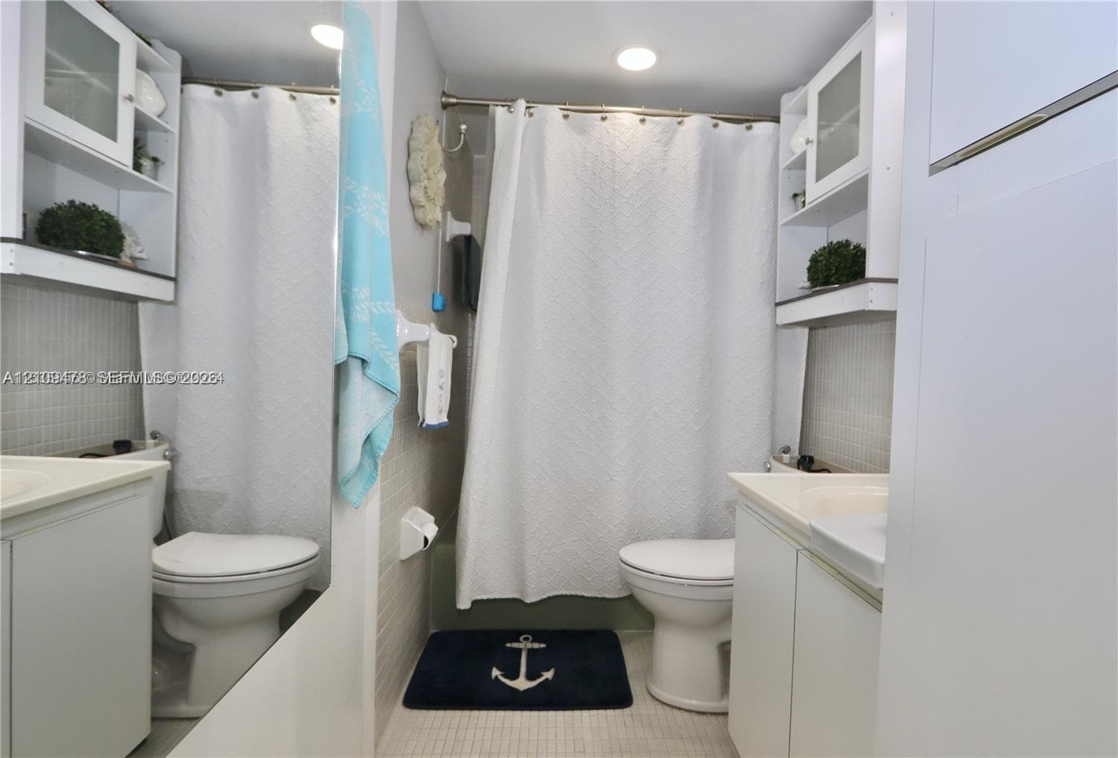 Full bathroom featuring dual white vanity cabinets, integrated white sinks, wall-mounted upper storage shelving, a tiled shower surround, and a large wall-mounted mirror
