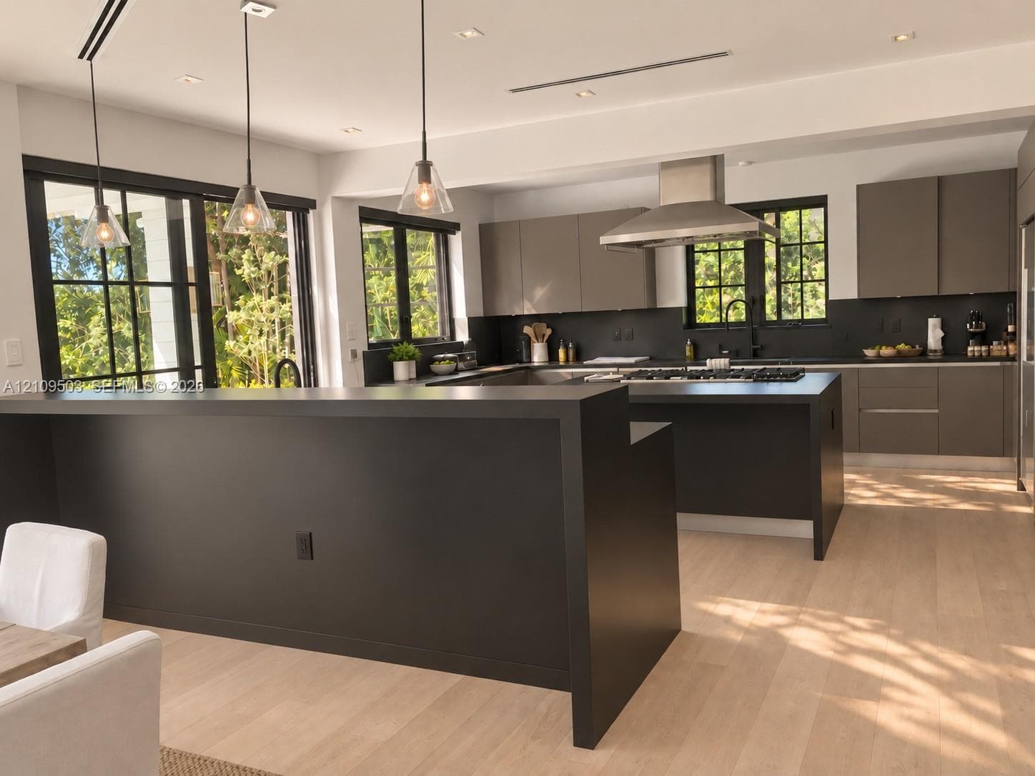 Kitchen featuring matte dark cabinetry, wood-finish flooring, a stainless steel range hood, glass pendant lighting, and a kitchen island with a dark countertop