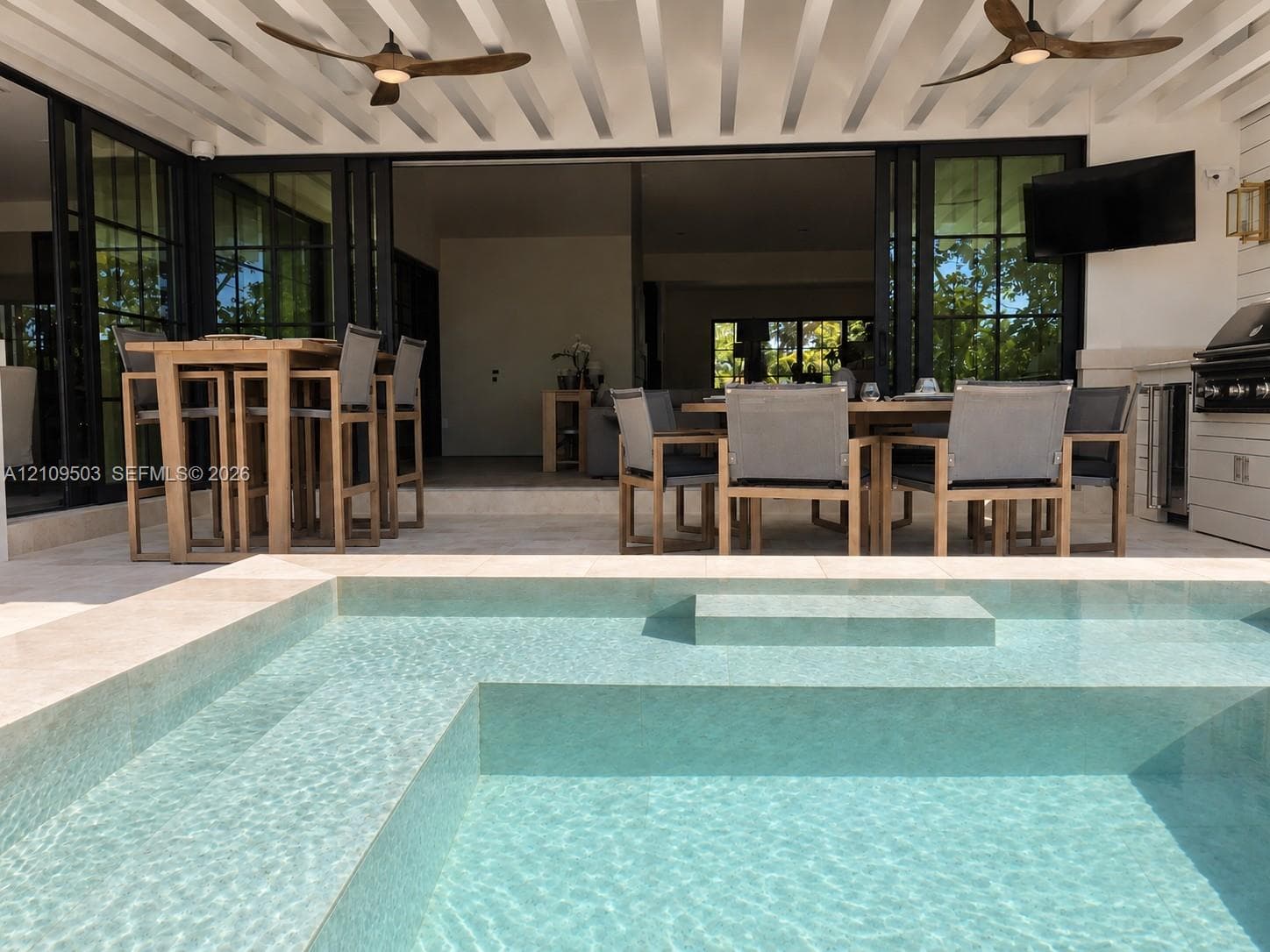 Covered patio featuring an integrated outdoor kitchen, multi-panel retractable glass doors, dual ceiling fans, and an inground swimming pool with stone-tiled decking