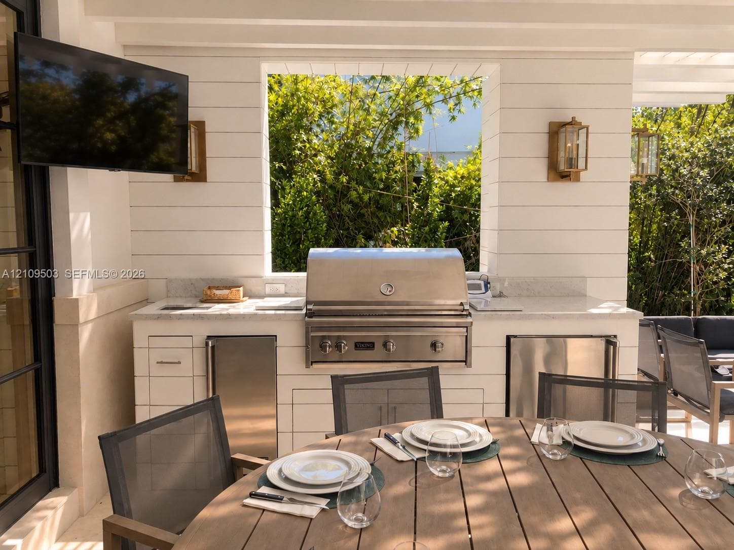 Covered outdoor kitchen featuring shiplap siding, built-in stainless steel grill, stone countertops, dual under-counter stainless steel appliances, and mounted lantern-style light fixtures
