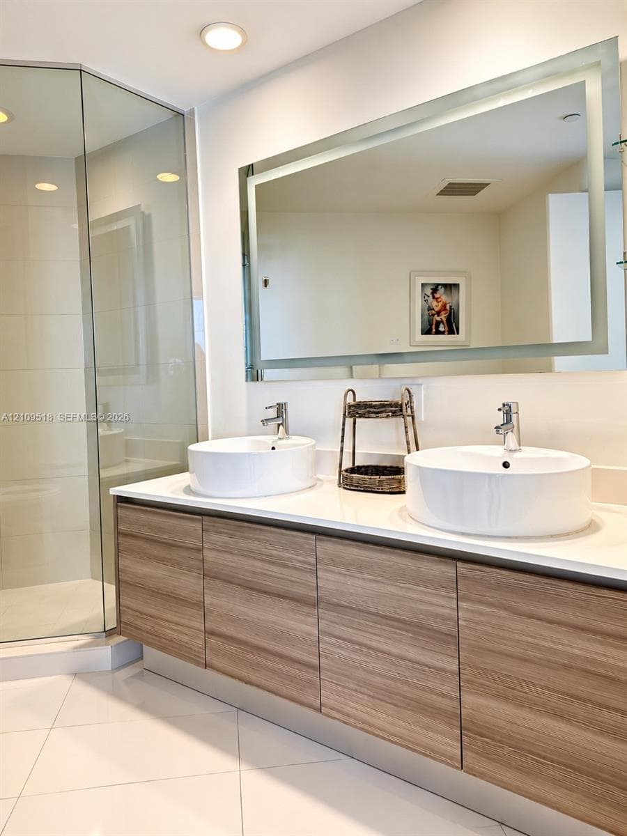 Floating vanity with wood-finish cabinetry, dual white vessel sinks, solid-surface countertop, and a large integrated LED-lit mirror, paired with a glass-enclosed shower featuring light-toned tile flooring