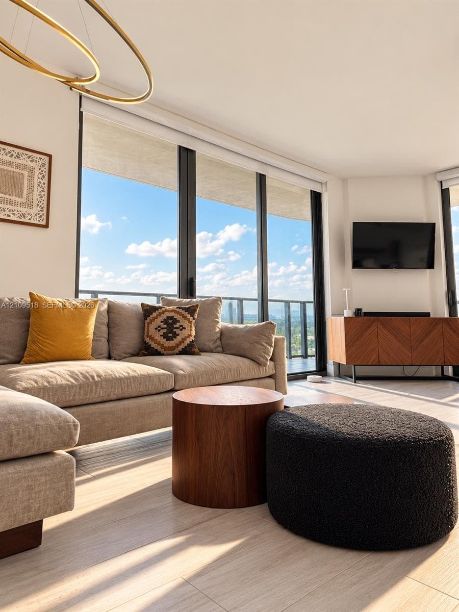 Floor-to-ceiling glass paneling with integrated balcony access, neutral stone-finish flooring, gold-tone modern circular chandelier, and custom built-in media cabinetry
