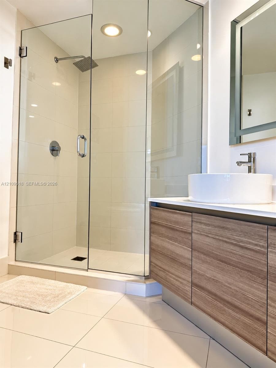 Floating wood-finish vanity with a white vessel sink, polished chrome faucet, glass-enclosed shower, and large-format tile flooring