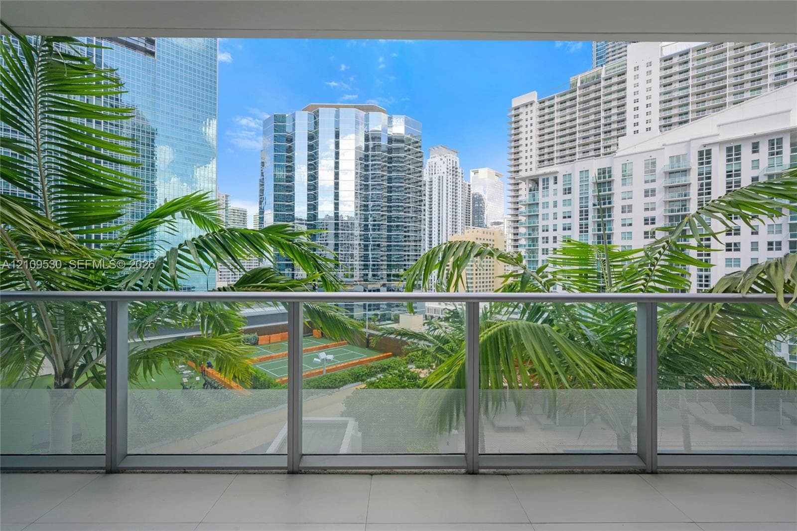 Covered balcony featuring tile flooring and a glass and metal railing, overlooking an outdoor tennis court with manicured palm trees