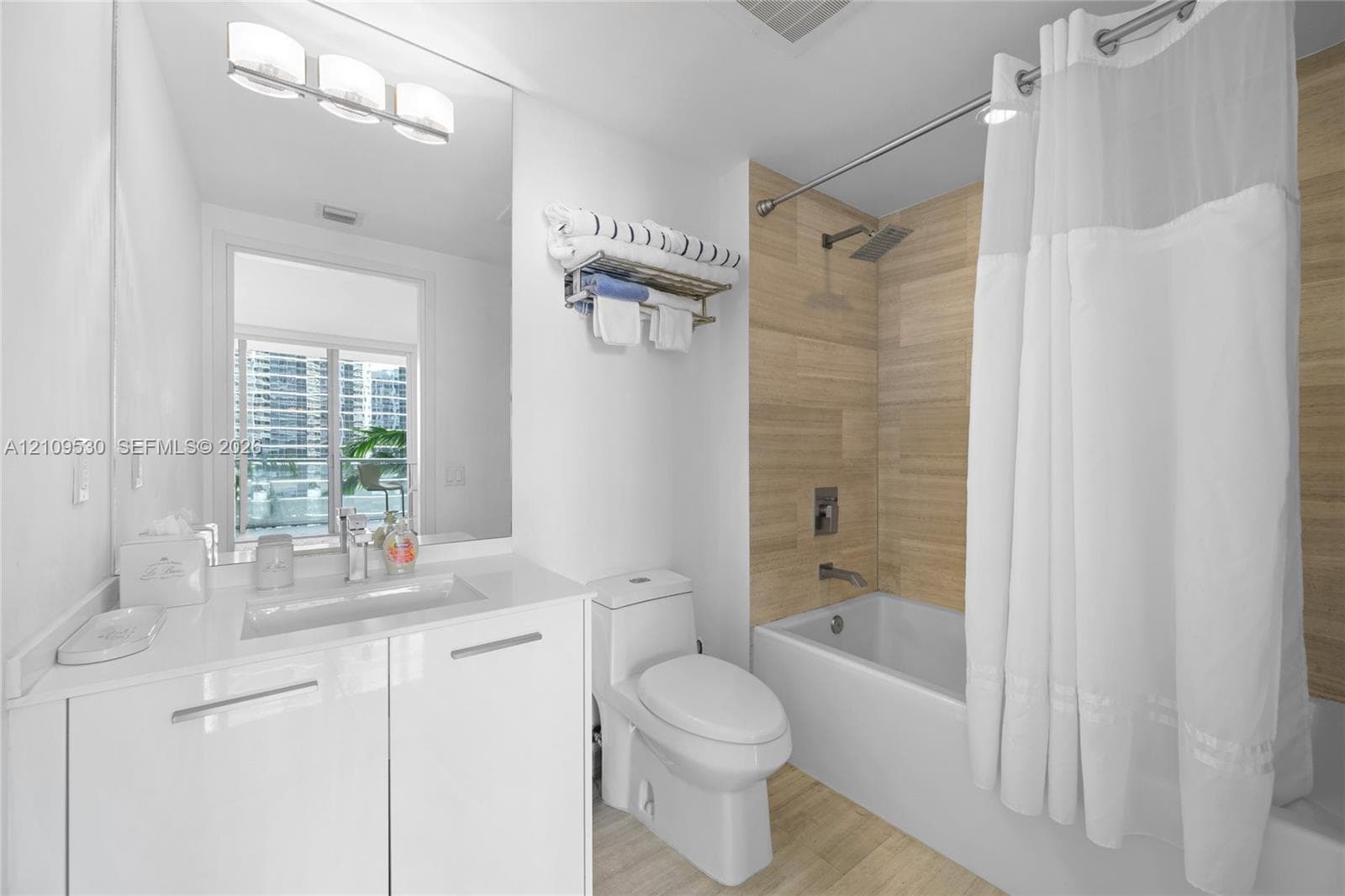 Bathroom featuring a white gloss vanity with an integrated sink, full-width mirror, wall-mounted light fixture, and a tub with wood-finish tile surround