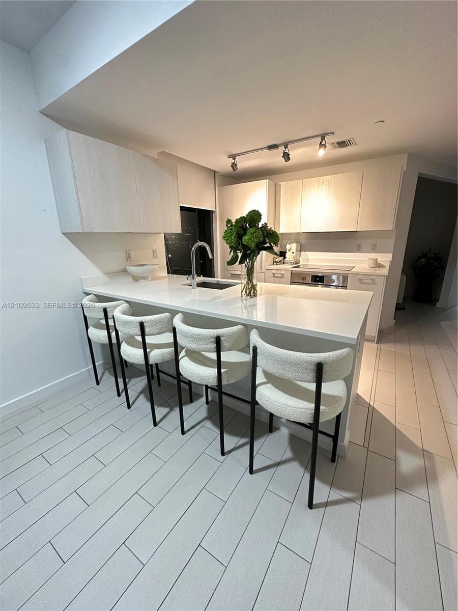 Kitchen featuring light-finish cabinetry, white solid-surface countertops, a center island with an undermount sink, built-in stainless steel appliances, and tile flooring
