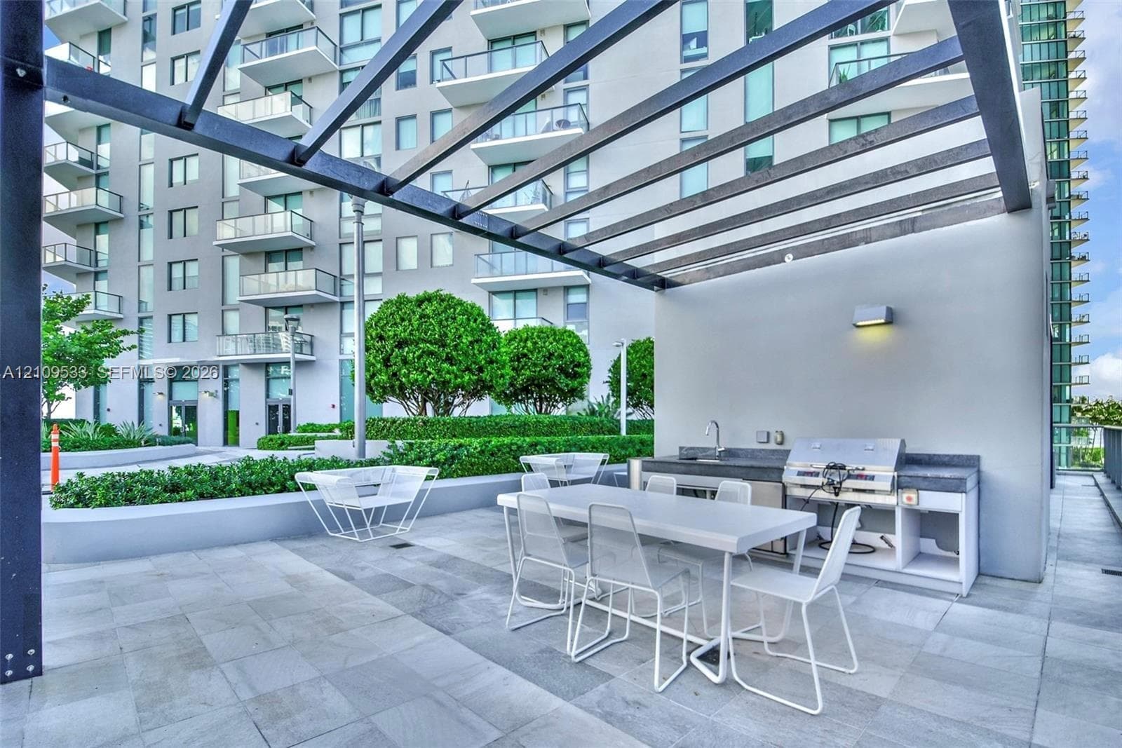 Outdoor terrace featuring an overhead pergola, built-in kitchen station with a stainless steel grill and sink, stone-finish flooring, and manicured landscaping
