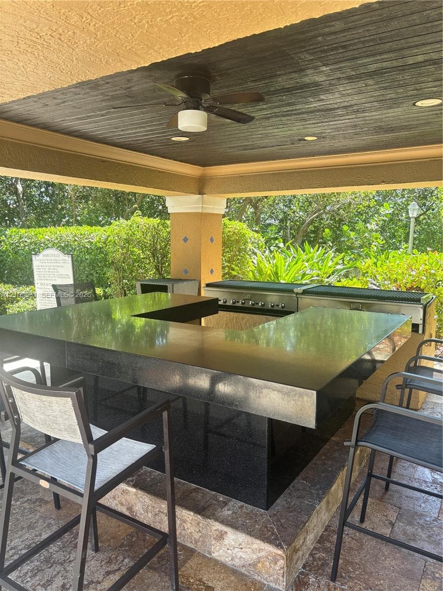 Covered outdoor kitchen featuring a stone-topped bar, stainless steel built-in grill, stone-tile flooring, and a ceiling fan