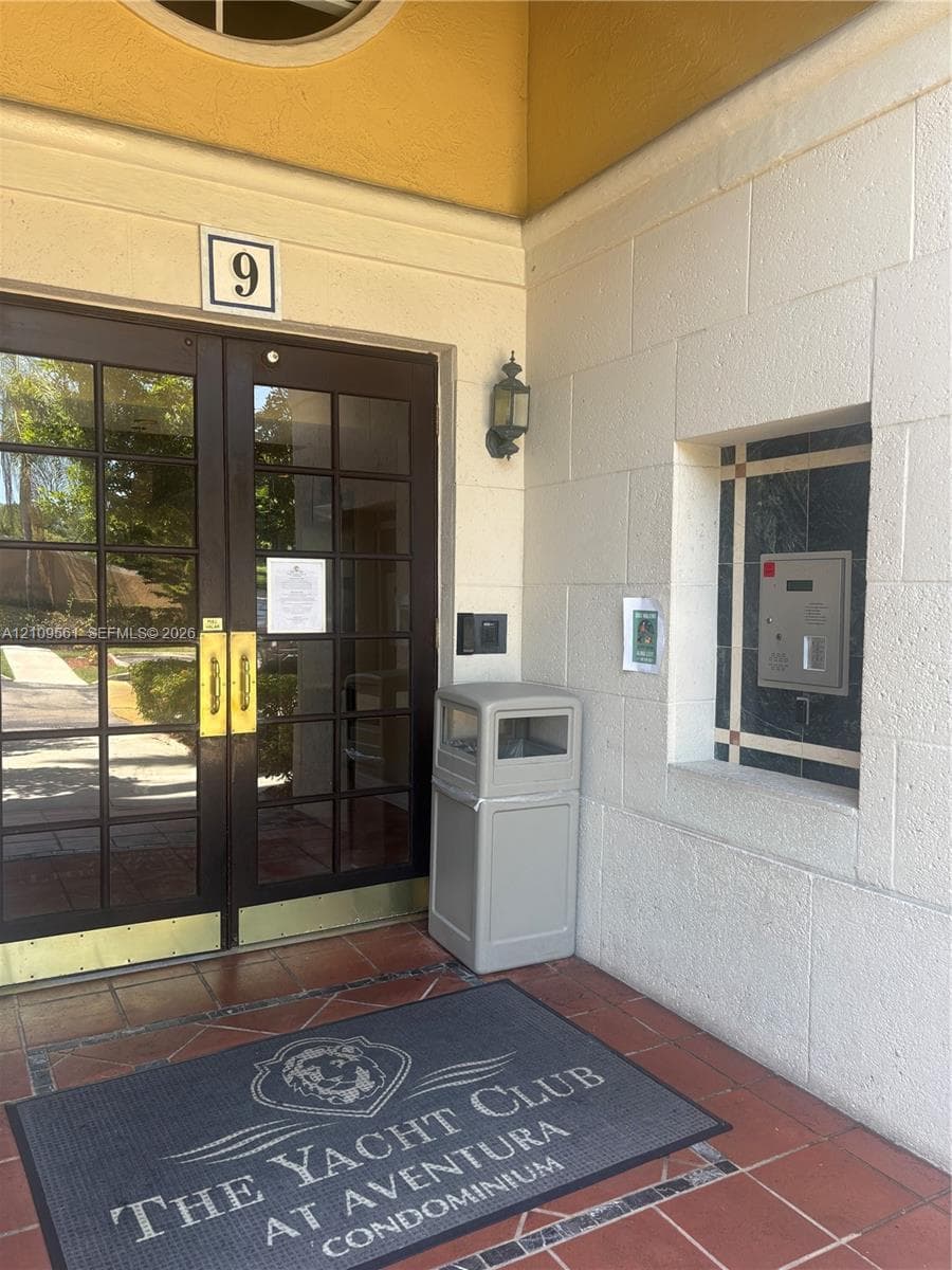 Building entrance featuring double glass doors with brass hardware, a security intercom system, a wall-mounted lantern-style light fixture, and terracotta tile flooring
