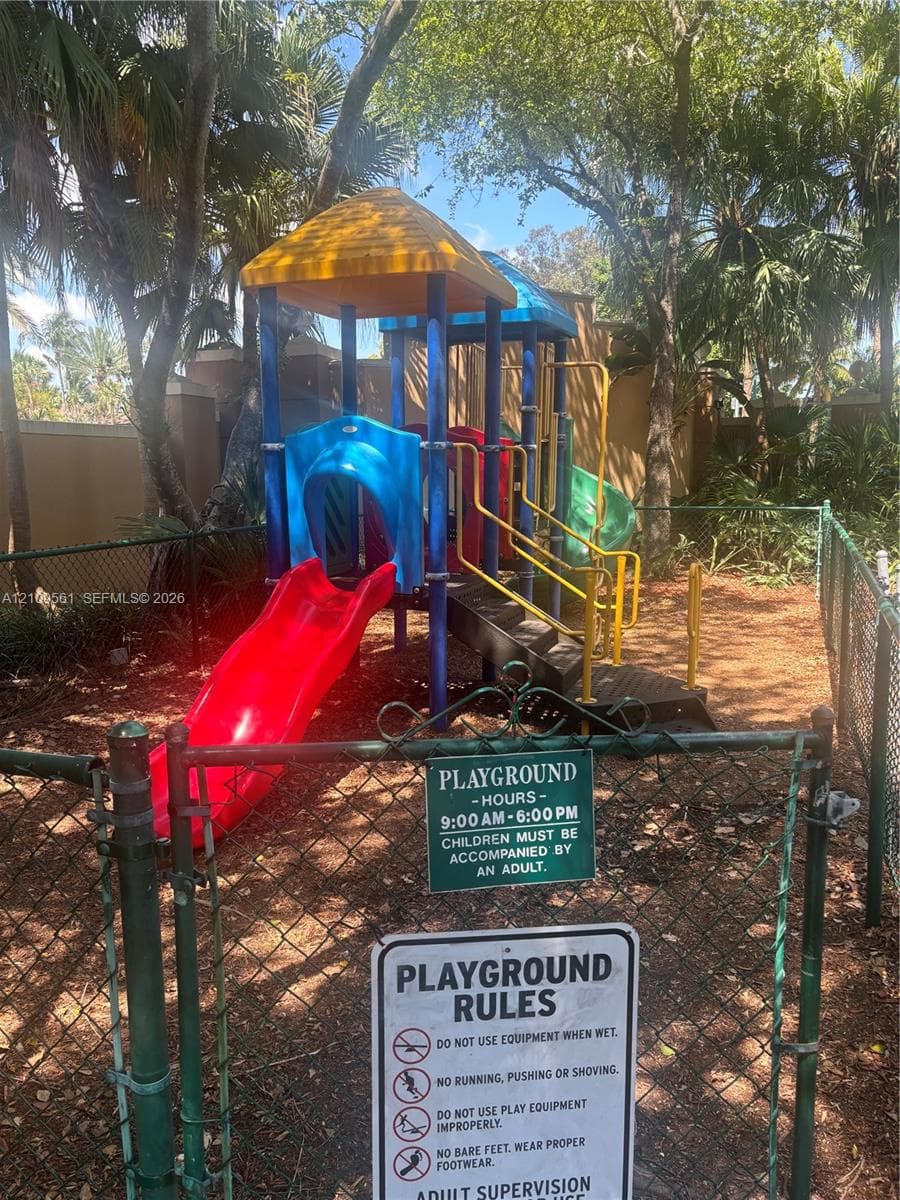 Enclosed recreation area featuring a multi-level climbing structure with slides, chain-link perimeter fencing, and mature canopy trees