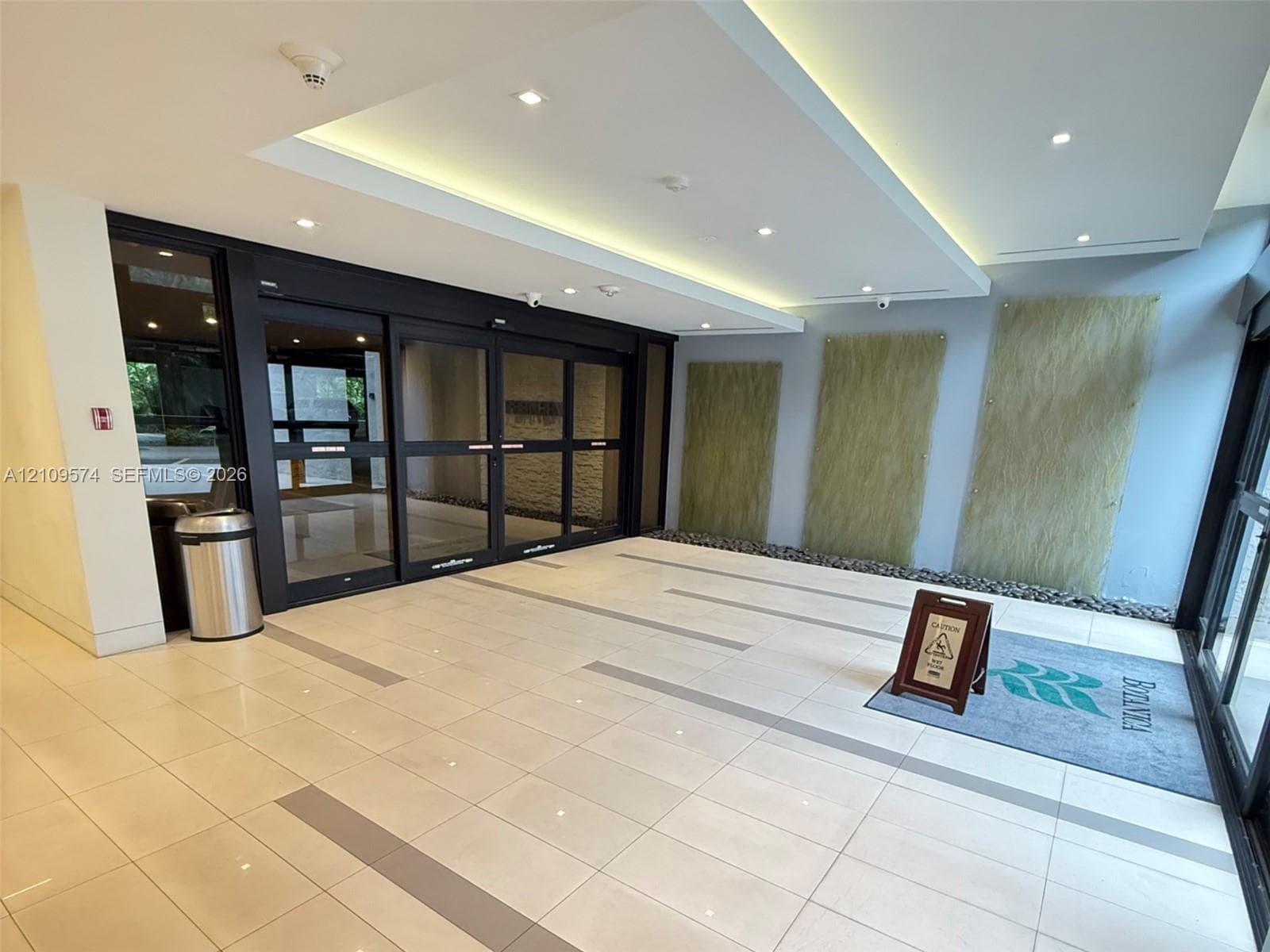 Lobby entrance featuring large-format tile flooring with linear accent inlays, automated sliding glass entry doors, recessed tray ceiling lighting, and textured wall paneling