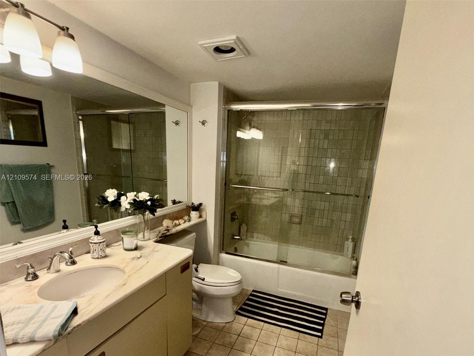 Full-width wall mirror, vanity cabinet with integrated sink, stone-finish countertop, shower and bathtub combination with sliding glass doors, and tiled flooring