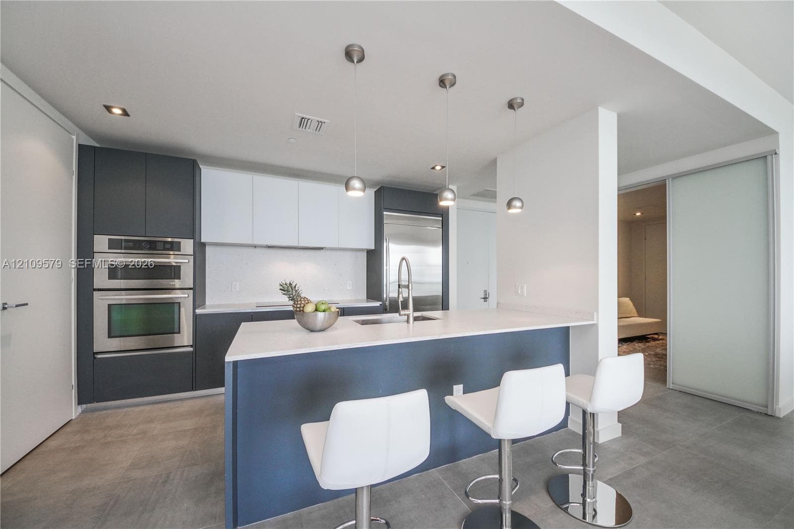 Modern kitchen featuring matte charcoal and white cabinetry, quartz countertops, stainless steel built-in double wall oven, central island with under-mount sink, and three pendant light fixtures